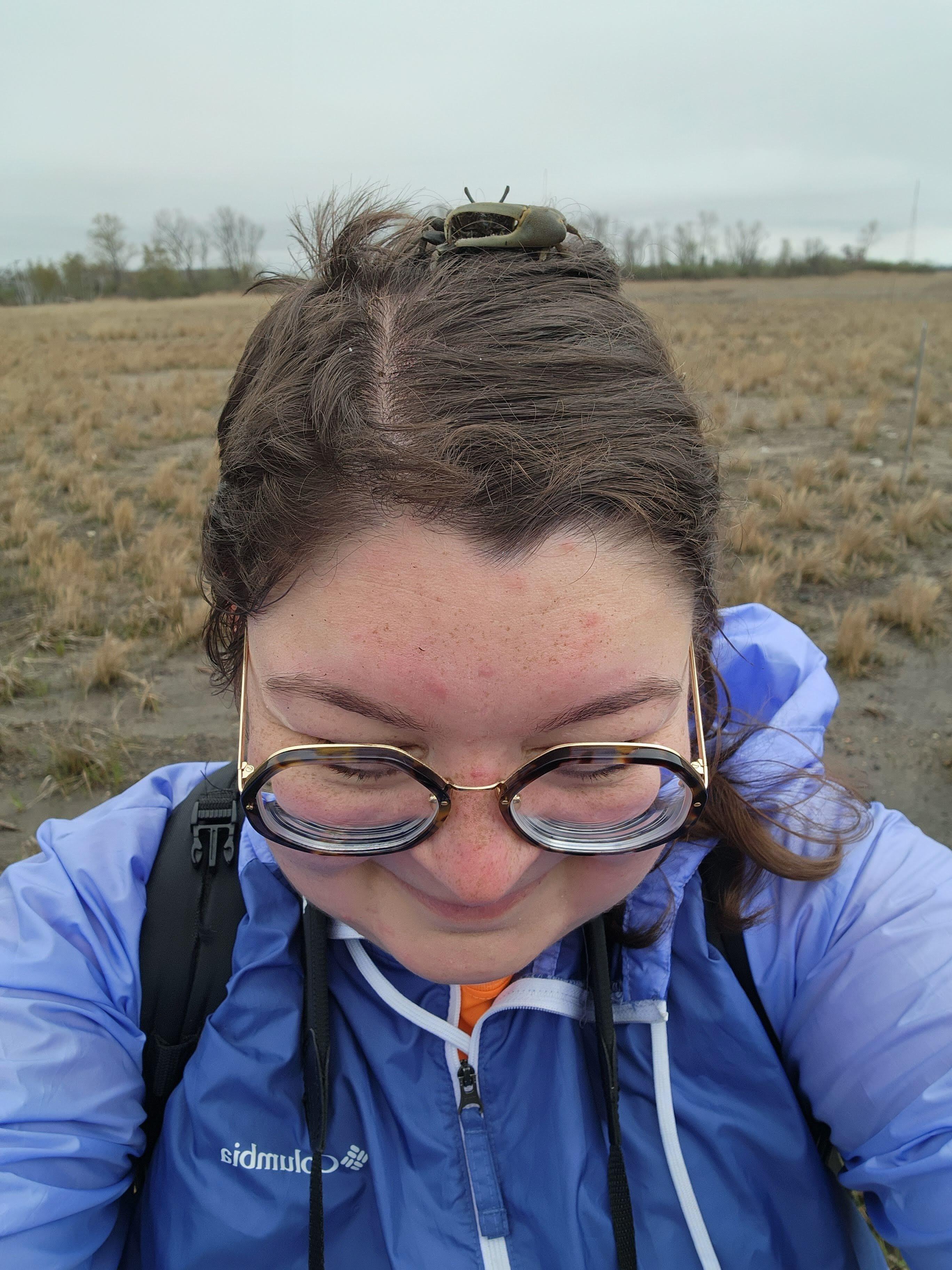 A selfie of Allison Middlemass out on the beach on a cloudy day, with a fiddler crab on her hair.