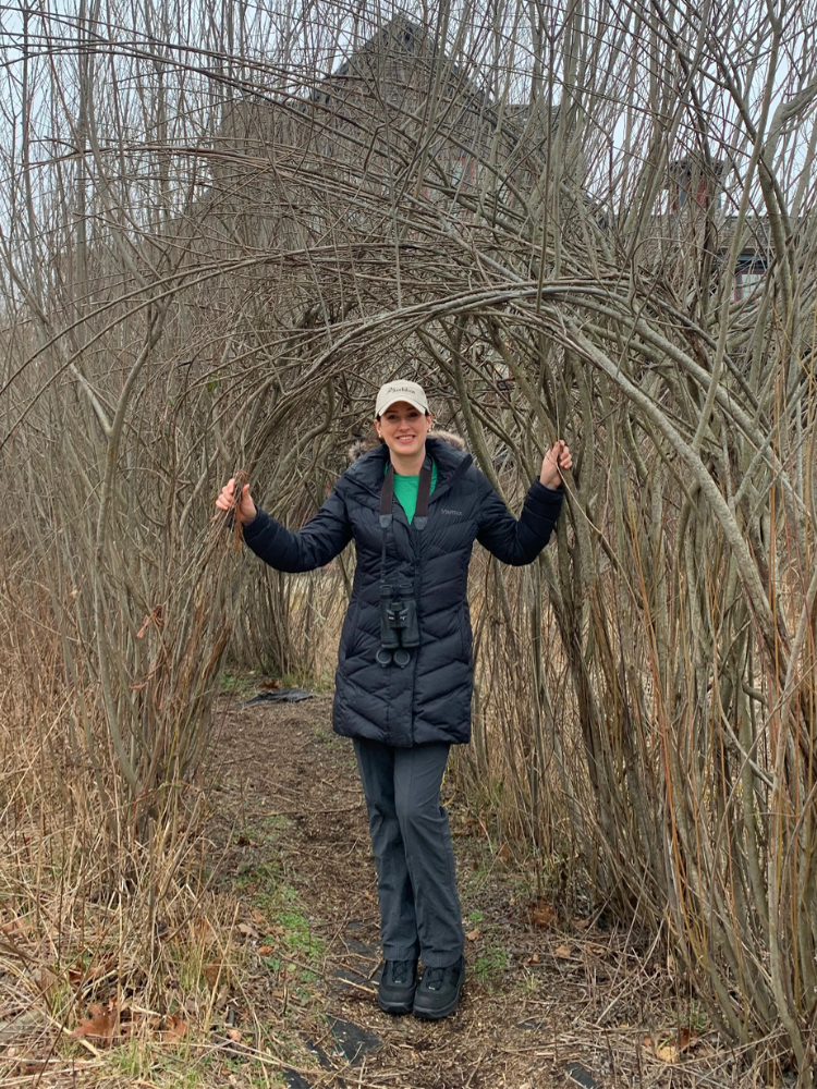 Rochelle Thomas stands in between a row of leafless shrubs in a long black puffer jacket. She is smiling.