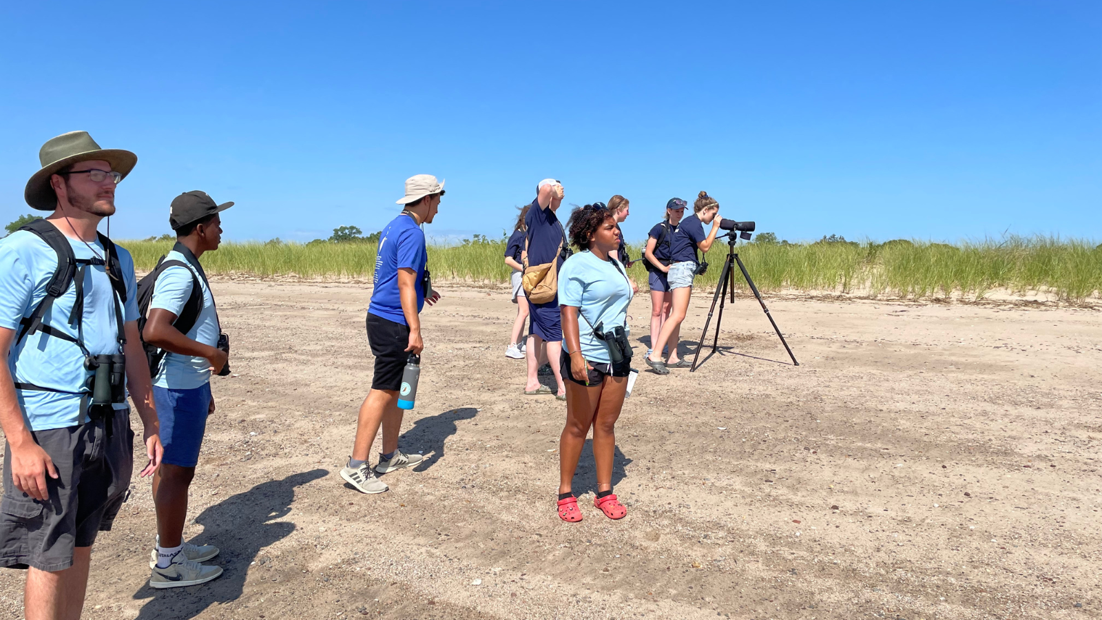 WildLife Guards and Junior Forest Technicians stand on a beach, spread out and using binoculars and guides to identify birds that are not visible in the photo.