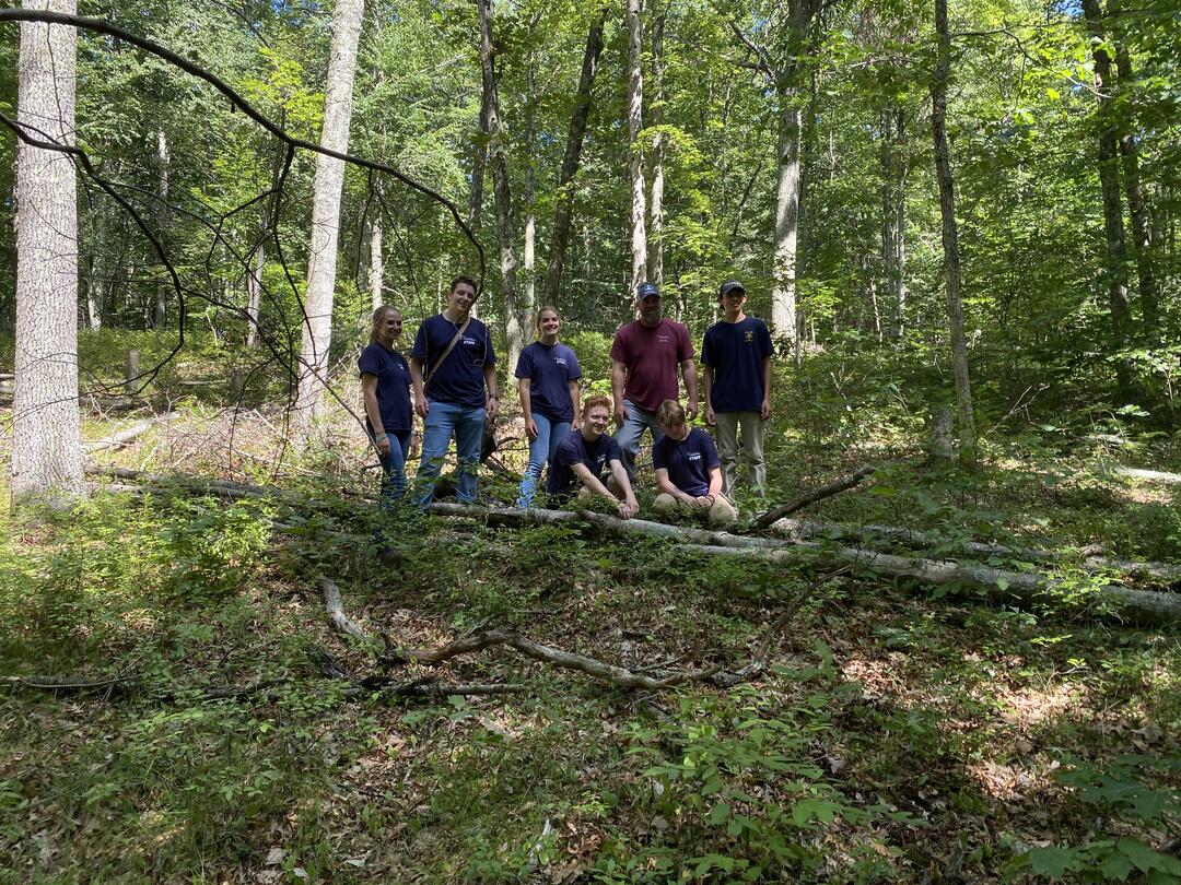 A group of Junior Forest Technicians and their mentor, Glen, pose for a group photo in a small wooded clearing.